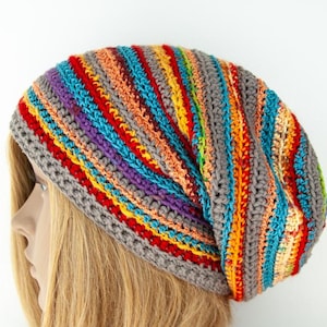 Op de afbeelding: Een handgemaakte, losse beanie in een levendige regenboog van kleuren. De hoed heeft strepen van rood, oranje, geel, groen, blauw en paars, allemaal tegen een grijze basis. Het haakwerk is zichtbaar en voegt textuur toe.