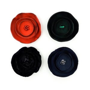 May include: Four handmade flower-shaped brooches in red, black, dark green, and navy blue. Each brooch has a layered petal design with a central embellishment. The brooches are set against a white background.