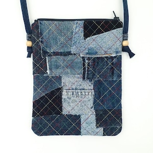 Op de afbeelding: Een crossbodytas van denim patchwork met een marineblauwe band en ritssluiting. De tas is gemaakt van verschillende stukken denim stof, aan elkaar gestikt met een ruitpatroon. Houten kralen accentueren de bandbevestiging. Ideaal voor het dragen van essentials.