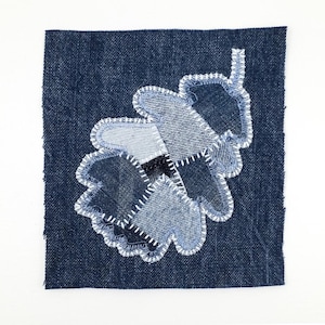 May include: A square dark blue denim fabric patch featuring an appliqued oak leaf design. The leaf is constructed from multiple shades of blue denim fabric scraps, joined together with visible white zigzag stitching along the edges.