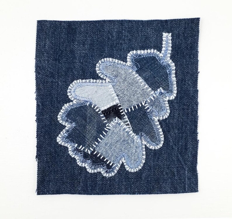 Peut inclure: Un carr&eacute; de tissu en jean bleu fonc&eacute; avec un motif de feuille de ch&ecirc;ne appliqu&eacute;. La feuille est compos&eacute;e de plusieurs morceaux de tissu en jean de diff&eacute;rentes nuances de bleu, assembl&eacute;s par des surpiq&ucirc;res blanches en zigzag visibles.