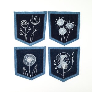 Può includere: Quattro tasche in tessuto denim blu scuro con ricami floreali bianchi. Ogni tasca ha un bordo azzurro con pois bianchi. I fiori ricamati includono una rosa, denti di leone e altri fiori stilizzati.