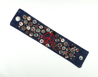 May include: Dark blue denim bracelet adorned with numerous gray and black buttons with red stitching. A red peace sign is embroidered in the center. Small beads and silver studs add detail to this handcrafted accessory.