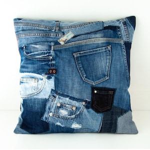 Può includere: Cuscino quadrato in denim riciclato. Il cuscino presenta un design patchwork con varie tonalità di denim blu, tra cui tasche e cuciture. Una piccola etichetta è visibile.