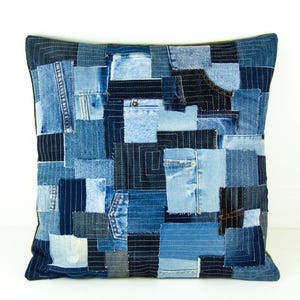 Puede incluir: Cojín cuadrado de patchwork de mezclilla azul. El diseño presenta una mezcla de piezas de mezclilla, incluyendo bolsillos y costuras, cosidas para crear un patrón texturizado único. Los bordes están acabados con un borde azul oscuro.