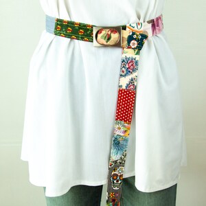 Op de afbeelding: Een kleurrijke patchwork stoffen riem met een zilveren gesp. De riem is gemaakt van verschillende stoffen met bloemen- en geometrische patronen. De riem is aan de voorkant in een knoop geknoopt.
