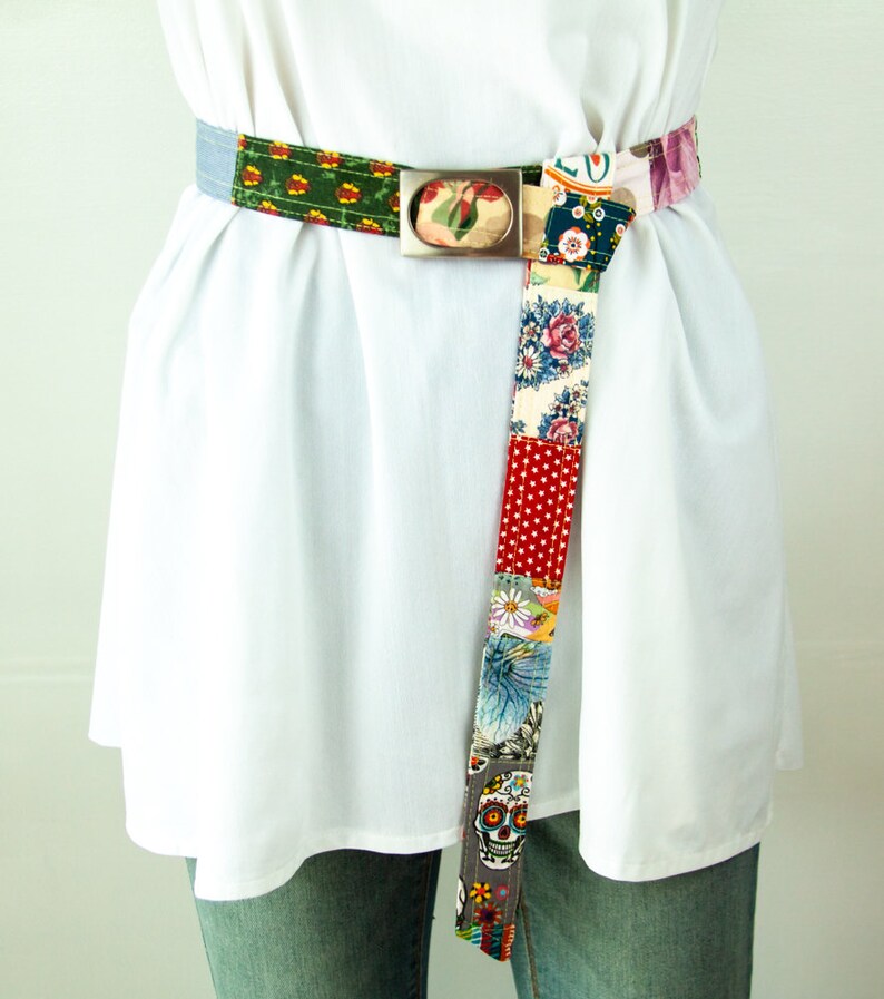 Op de afbeelding: Een kleurrijke patchwork stoffen riem met een zilveren gesp. De riem is gemaakt van verschillende stoffen met bloemen- en geometrische patronen. De riem is aan de voorkant in een knoop geknoopt.