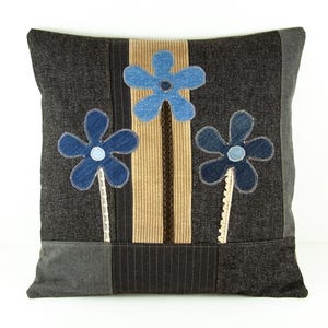 Può includere: Cuscino decorativo quadrato con sfondo grigio scuro e tre applicazioni di fiori in denim blu. I fiori hanno centri bianchi e sono posizionati su un pannello a righe beige e marrone.