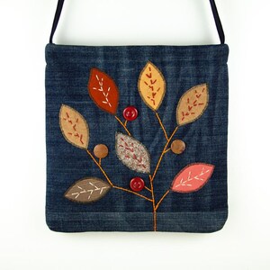 Op de afbeelding: Denim crossbody tas met een gestikt bladerontwerp. De bladeren zijn in tinten bruin, oranje en geel, met rode knoopaccenten. De tas heeft een zwarte band en een vierkante vorm.