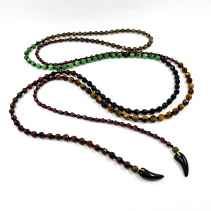 May include: A layered necklace with four strands of beads. The strands are made of brown, green, black, and red beads. The necklace has two black horn-shaped pendants.