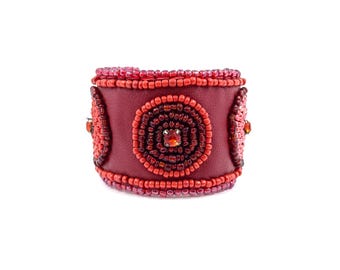 May include: A wide, burgundy leather cuff bracelet. The bracelet is adorned with red and pink beads, forming concentric circles and borders. A central, square-cut, orange-red gemstone is set in the center of the design. Additional gemstones are set on the sides.