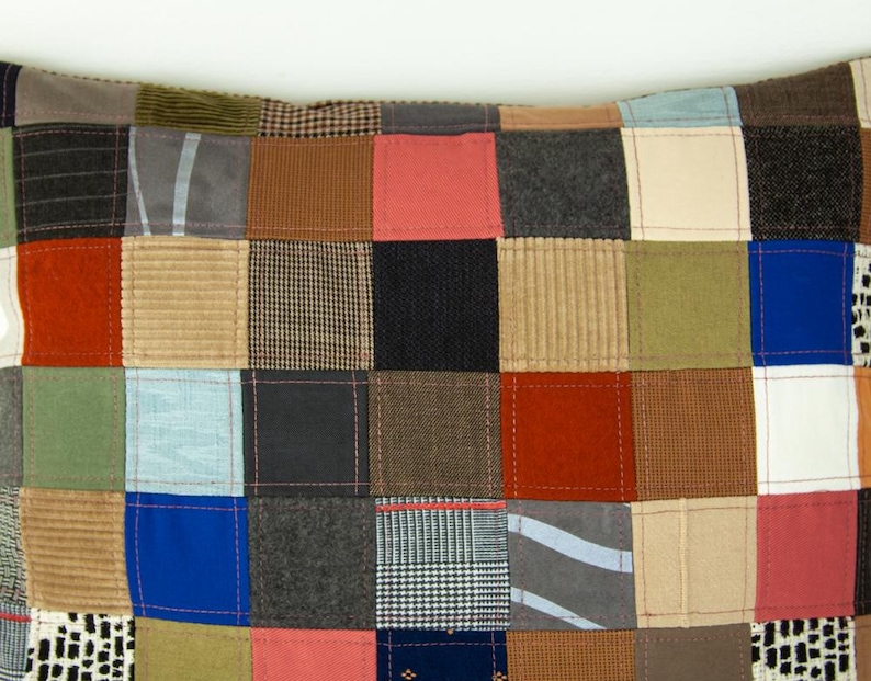 Puede incluir: Un coj&iacute;n patchwork con una variedad de cuadrados de tela en tonos de rojo, azul, verde, marr&oacute;n y gris. Los cuadrados est&aacute;n cosidos, creando una superficie texturizada. El dise&ntilde;o es una mezcla de patrones y texturas.