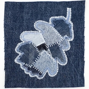 Peut inclure: Un carr&eacute; de tissu en jean bleu fonc&eacute; pr&eacute;sentant une applique en forme de feuille de ch&ecirc;ne. La feuille est compos&eacute;e de plusieurs pi&egrave;ces de jean dans des nuances de bleu, fix&eacute;es par des points de zigzag blancs sur les bords de chaque section.