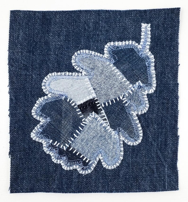 Peut inclure: Un carr&eacute; de tissu en jean bleu fonc&eacute; pr&eacute;sentant une applique en forme de feuille de ch&ecirc;ne. La feuille est compos&eacute;e de plusieurs pi&egrave;ces de jean dans des nuances de bleu, fix&eacute;es par des points de zigzag blancs sur les bords de chaque section.