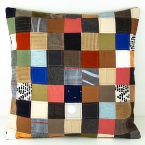Puede incluir: Coj&iacute;n cuadrado de patchwork con una variedad de cuadrados de tela en tonos marrones, azules, rojos y verdes. El coj&iacute;n tiene un fondo neutro y est&aacute; hecho de varias texturas, incluyendo pana y telas estampadas. El coj&iacute;n es cuadrado.