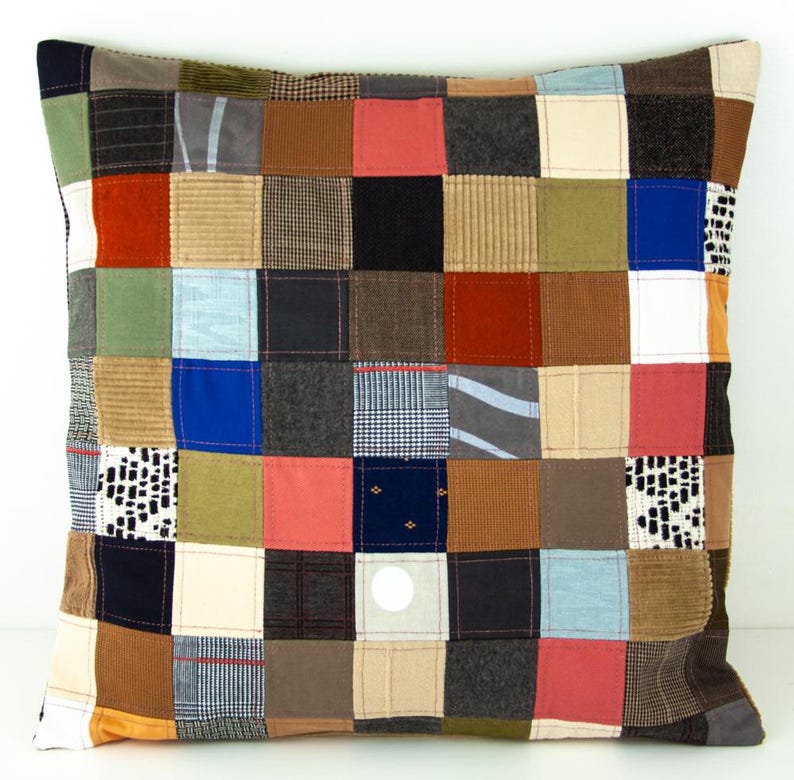 Puede incluir: Coj&iacute;n cuadrado de patchwork con una variedad de cuadrados de tela en tonos marrones, azules, rojos y verdes. El coj&iacute;n tiene un fondo neutro y est&aacute; hecho de varias texturas, incluyendo pana y telas estampadas. El coj&iacute;n es cuadrado.