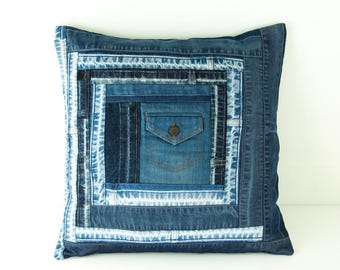 May include: Square denim pillow with a patchwork design. The pillow features various shades of blue denim, including a pocket detail with a button closure. The design incorporates different textures and patterns, creating a unique and rustic aesthetic.