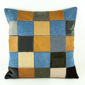 May include: Square patchwork pillow with a variety of textures and colors. The design features squares of denim blue, tan leather, and various other fabrics. The pillow is a decorative home accent.