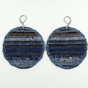 May include: A pair of circular earrings crafted from layered blue denim fabric. Each earring features horizontal stripes of varying shades of blue, accented with intricate decorative stitching in white, gold, and dark thread, finished with silver-tone leverback hooks.