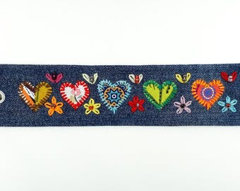 May include: A denim bracelet with colorful embroidered hearts and floral accents. The bracelet features a variety of heart designs in yellow, red, blue, and orange, with embroidered flowers and butterflies. Fastened with a silver snap.