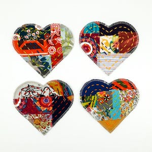 Op de afbeelding: Vier hartvormige stoffen onderzetters met een patchworkontwerp van diverse kleurrijke patronen, waaronder bloemmotieven, geometrische en abstracte prints. Elke onderzetter is afgewerkt met zichtbaar decoratief handborduurwerk.