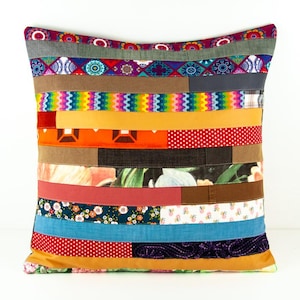 Peut inclure: Un coussin carré décoratif avec un motif patchwork. Le coussin présente des rayures horizontales de différentes couleurs et motifs, notamment floraux, à pois et géométriques. La palette de couleurs comprend le rouge, l'orange, le bleu et le vert.