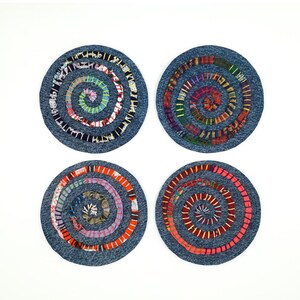 May include: Four circular coasters made of blue denim fabric. Each coaster features a spiral design created with colorful, multi-patterned fabric strips stitched onto the surface. The spirals vary in color, including shades of red, orange, yellow, green, purple, and white, creating a distinct, textured appearance on each individual denim base.