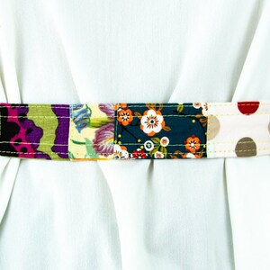 Op de afbeelding: Een kleurrijke patchwork stoffen riem met verschillende bloemen- en geometrische patronen in tinten blauw, groen, rood, roze, geel en bruin. De riem is met een lichtgele draad aan elkaar genaaid.