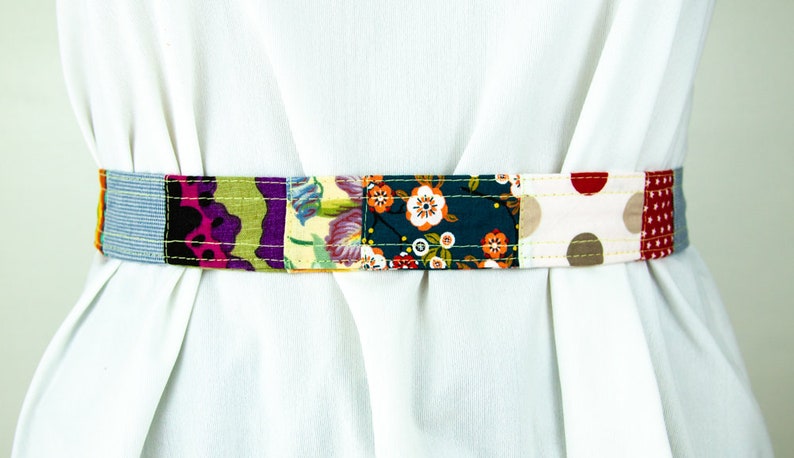 Op de afbeelding: Een kleurrijke patchwork stoffen riem met verschillende bloemen- en geometrische patronen in tinten blauw, groen, rood, roze, geel en bruin. De riem is met een lichtgele draad aan elkaar genaaid.
