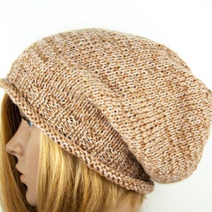 Può includere: Un berretto lavorato a maglia nei toni del beige e del marrone. Il cappello ha un bordo ripiegato e una vestibilità ampia. Il motivo a maglia è visibile, mostrando la trama del filato. Il cappello è mostrato su una testa di manichino con capelli biondi.