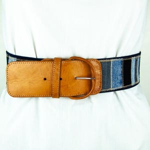 Op de afbeelding: Een brede riem met een lichtbruine leren gesp en riem. De body van de riem is gemaakt van denimstof in tinten blauw en zwart. De riem wordt over een wit kledingstuk gedragen.