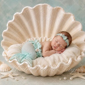 Op de afbeelding: Een pasgeboren baby slaapt in een grote, crèmekleurige decoratieve schelp. De baby draagt een lichtblauwe gehaakte zeemeerminstaart en een bijpassende hoofdband met kleine witte parels en schelpjes, liggend op een zacht wit kussentje.