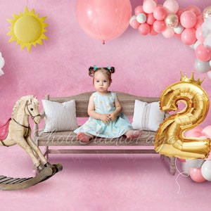 May include: A little girl sits on a wooden bench with a rocking horse and a gold foil number two balloon. The background is pink with a cloud, a sun, and a balloon arch.
