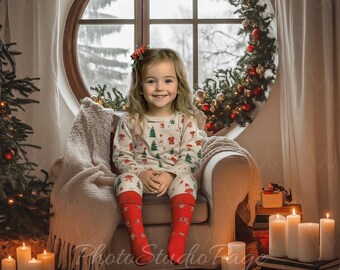 May include: A young girl wearing a white and green Christmas pajamas sits in a cream-colored armchair in front of a window with a garland of greenery and red ornaments. The room is decorated with a Christmas tree and lit candles.