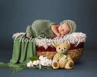 May include: A newborn baby sleeping in a wicker basket lined with a white faux fur blanket. The baby is wrapped in a green knitted blanket and wearing a white crocheted hat. A green blanket is draped over the side of the basket. A brown teddy bear with a green ribbon is sitting in front of the basket. White flowers are in the foreground.
