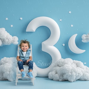May include: A young child sits on a white chair in front of a large white number three. The background is a light blue wall with white clouds and stars.