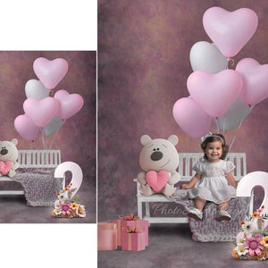 May include: A little girl sits on a white bench with a teddy bear holding a pink heart. The bench is decorated with a pink and white knitted blanket and a floral arrangement. There are pink heart-shaped balloons in the background.