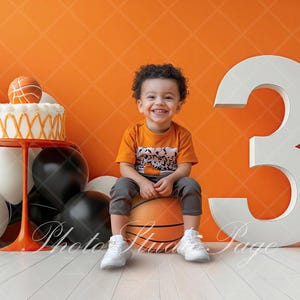 Puede incluir: Un niño sonriente está sentado en una pelota de baloncesto, rodeado de globos y un pastel. El pastel está cubierto con una pelota de baloncesto y está sobre un soporte rojo. Un gran número 3 blanco está detrás del niño. El fondo es naranja.