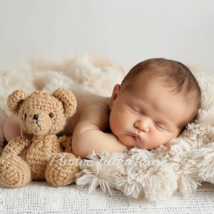 May include: A newborn baby sleeping peacefully on a white fluffy blanket with a crocheted teddy bear beside them.