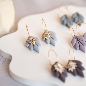 Op de afbeelding: Een set van zes paar oorbellen met een bladerenontwerp en een kristalaccent. De oorbellen zijn gemaakt van goudkleurig metaal en hebben een hoepelvorm. De bladeren zijn in grijstinten en paars.