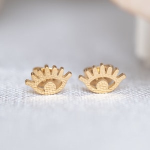 Op de afbeelding: Een paar goudkleurige oorstekers in de vorm van gestileerde ogen. De oorbellen hebben een textuur en staan tegen een zachte, neutrale achtergrond. Het ontwerp heeft een gedetailleerde wimper en iris.