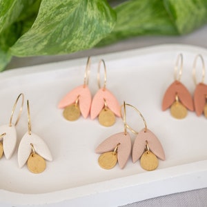 Könnte beinhalten: Drei Paar Ohrringe aus Ton mit goldenen Kreisen und Anhängern. Die Ohrringe sind in den Farben Rosa, Pfirsich und Weiß.