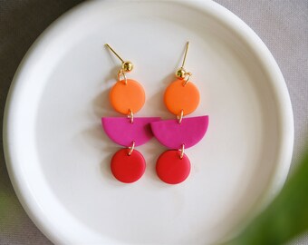May include: A pair of dangle earrings with three geometric shapes. The top shape is a circle in orange, the middle shape is a half-circle in pink, and the bottom shape is a circle in red.