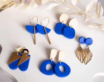 May include: Five pairs of blue and gold earrings. The earrings are made of polymer clay and feature different shapes, including circles, ovals, and triangles. Some of the earrings have gold accents.