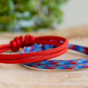 Pu&ograve; includere: Due braccialetti in corda intrecciata, uno rosso e uno blu con strisce rosse e bianche. I braccialetti sono su una superficie di legno.