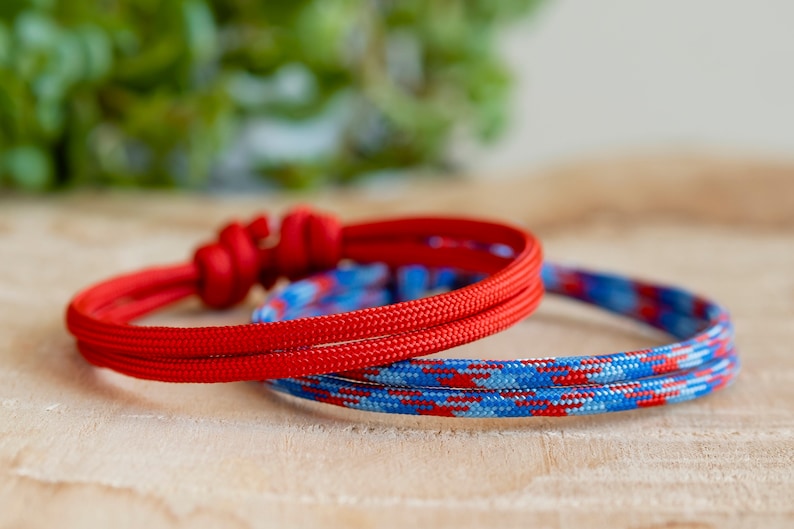 Pu&ograve; includere: Due braccialetti in corda intrecciata, uno rosso e uno blu con strisce rosse e bianche. I braccialetti sono su una superficie di legno.
