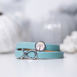 Può includere: Un bracciale avvolgente in pelle azzurra con un ciondolo argentato a forma di pesce e un ciondolo circolare a cabochon che mostra l'illustrazione di un albero con vari piccoli cerchi colorati tra i rami su sfondo bianco.