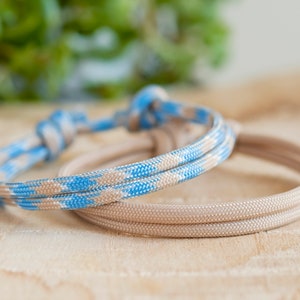Pu&ograve; includere: Due braccialetti in corda intrecciata, uno beige e uno blu e beige. I braccialetti sono annodati e sono su una superficie di legno chiaro.