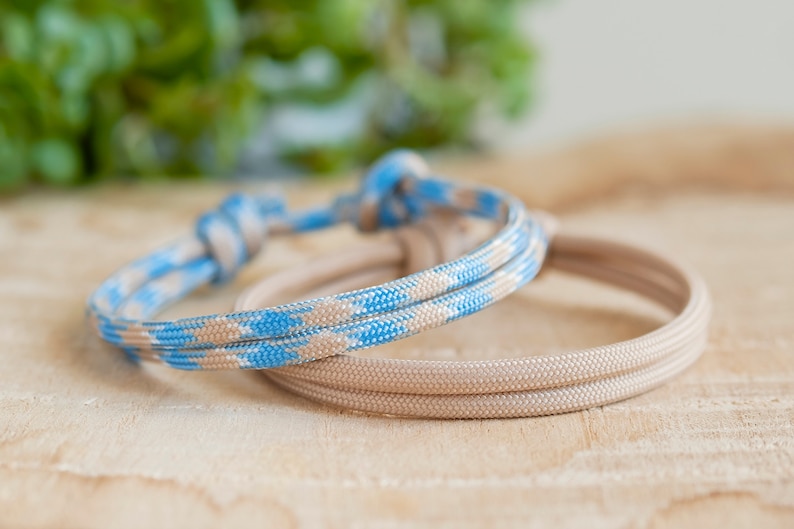 Pu&ograve; includere: Due braccialetti in corda intrecciata, uno beige e uno blu e beige. I braccialetti sono annodati e sono su una superficie di legno chiaro.