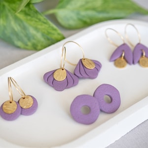 May include: A selection of handmade earrings in different styles, presented on a white tray. The earrings are a soft purple with gold-coloured details. Designs include hoop and stud earrings.